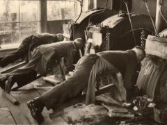 Historisches Foto der Edelsteinschleiferei. Drei Männer schleifen im liegen Edelsteine an einer Maschine.